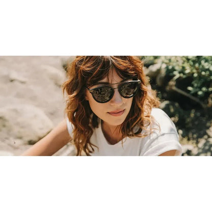 A woman wearing Shwood Kinsrow sunglasses poses outdoors with a rocky background.