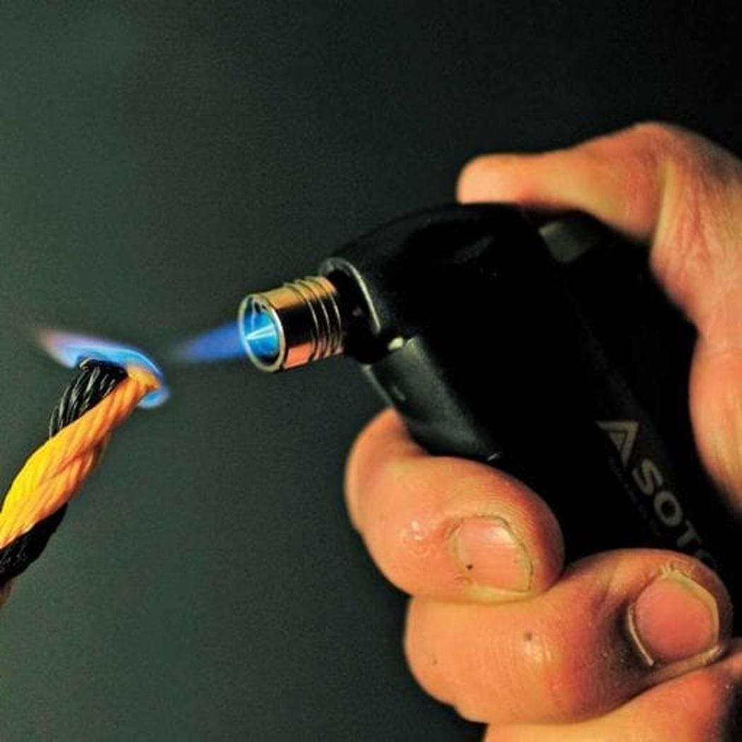 A hand holds the Soto Outdoors Pocket Torch with a blue flame aimed at a yellow and black rope against a dark background.