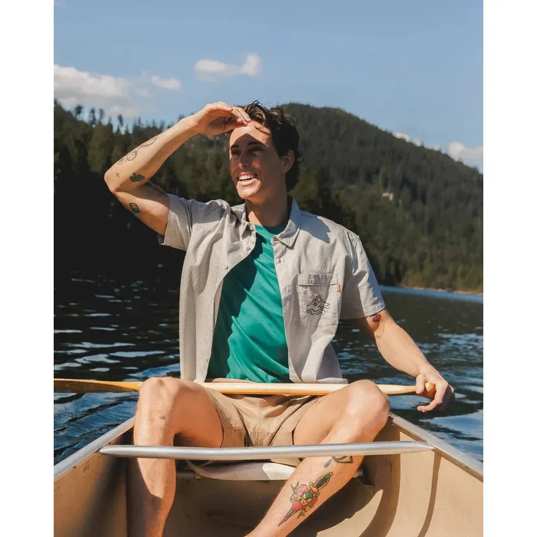 A man in a Tentree Men's Hopkins Shirt sits in a boat on a lake with mountains in the background.