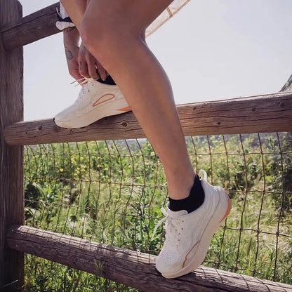 A woman adjusts her Teva Women's Trailwinder Low in Birch/ Peach Bloom while standing on a wooden fence with greenery in the background.