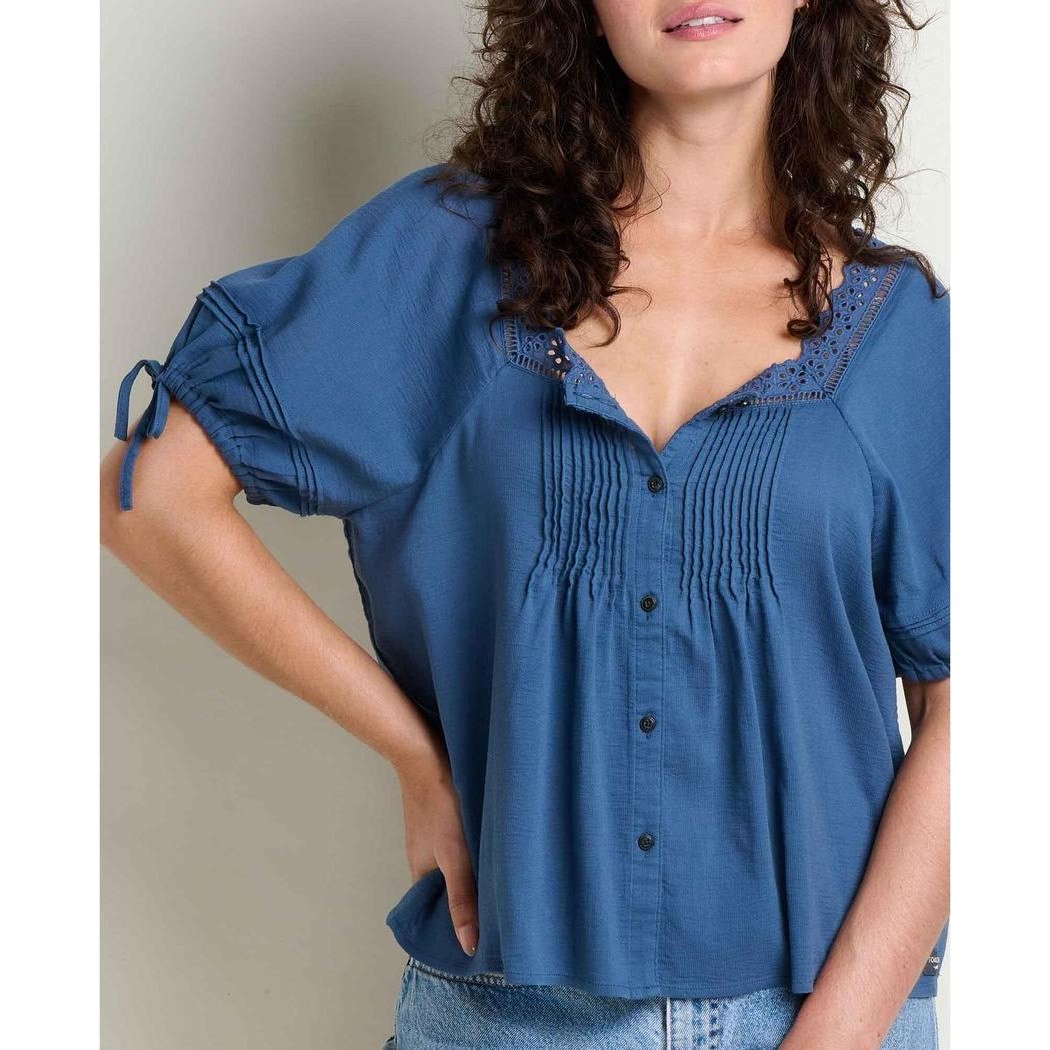 A woman wears the Toad & Co Women's Manzana Peasant Shirt in Canal blue with a V-neck and button front, against a plain background.