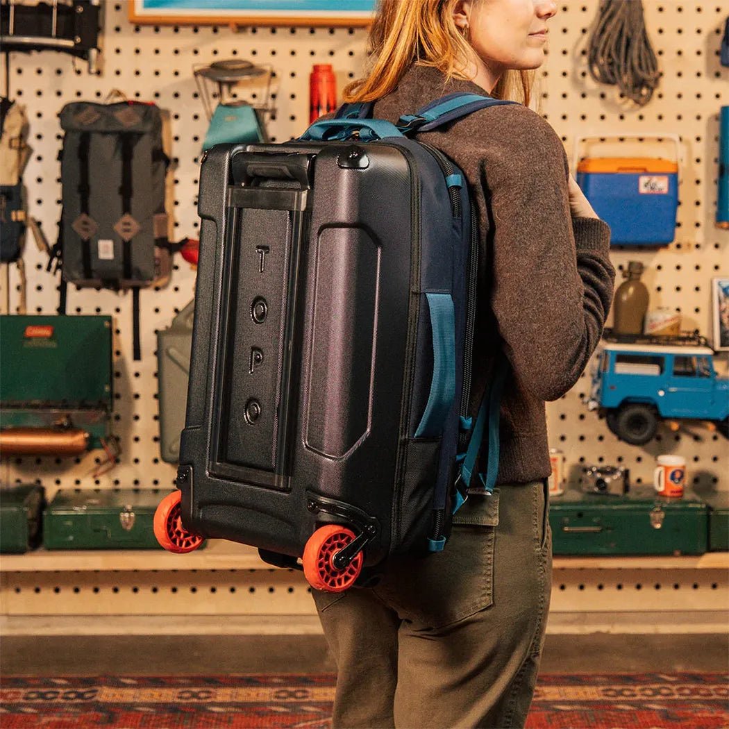 A woman wears the Topo Designs Global Travel Bag Roller in Navy/Sahara with orange wheels, standing in front of a pegboard wall with outdoor gear.