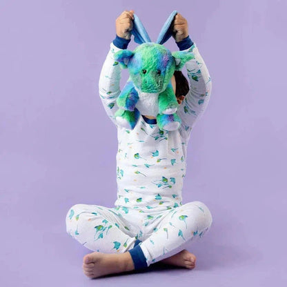 A child sits cross-legged on a purple background wearing white pajamas with green leaves, holding a blue and green Warmies Baby Dragon plush above their head.